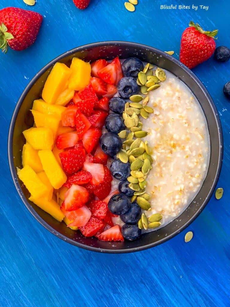 Steel cut oatmeal in a black bowl with mango, strawberry, blueberry and pumpkin seeds.