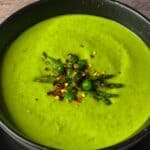 Peas and asparagus soup in a black bowl with roasted asparagus, peas and chilli flakes on top.
