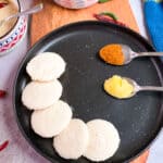 5 Idli on a black plate with a spice powder and ghee in spoons.