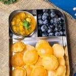 Puri in small circle shapes with Kale Chana Masala and blueberries in a lunchbox, kale in the background.