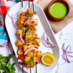 Tofu tikka on skewers on a white serving plate with Green mint chutney on the side.