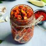 Instant mango pickle in a mason jar with curry leaves, red chili powder, garlic and raw mango pieces around it.