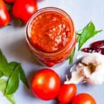 Tomato pickle in a mason jar with garlic, curry leaves and tomatoes around it.