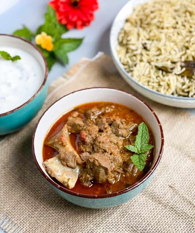 mutton curry in a bowl with mint leaf on top and mint pulao and onion raita in the back.